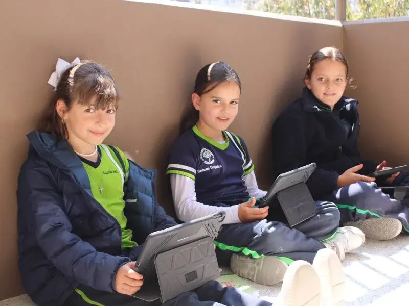 Tres estudiantes sentadas utilizan tabletas digitales en una actividad colaborativa al aire libre en el colegio.