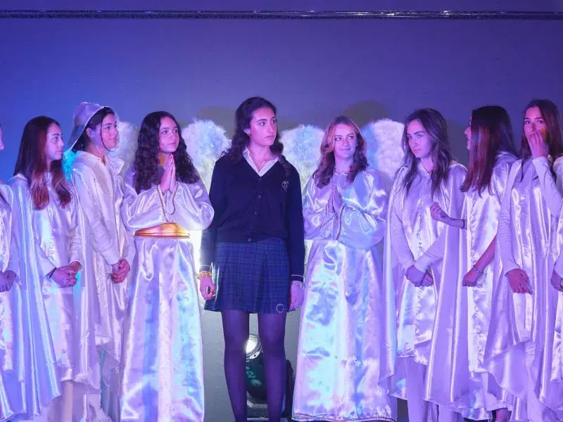Grupo de estudiantes en escena con alas blancas iluminadas participando en una presentación teatral escolar